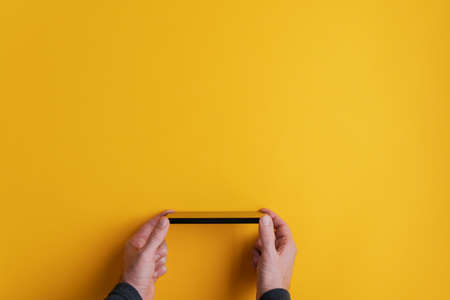 Male hands holding his black smart phone horizontally. Over yellow background with copy space ready fro your text or symbol. の写真素材