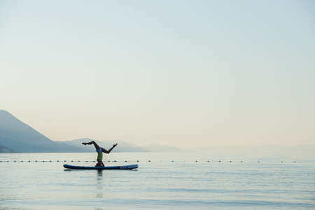 Fit young woman making a head stand on sup board floating on the calm water of morning sea.