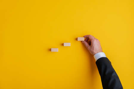 Hand of a businessman positioning wooden blocks in a stairway like structure in a conceptual image of business vision, strategy and promotion. Over yellow background with copy space.の写真素材