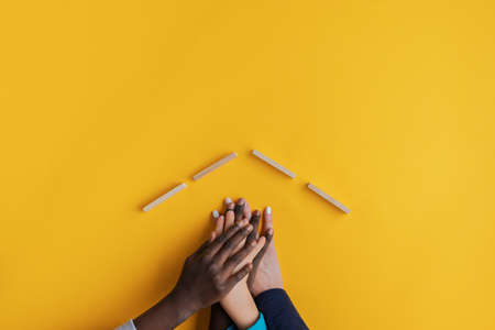 Hands of a mother and three children stacked under a roof made of wooden blocks. In a conceptual image over yellow background with copy space.の写真素材