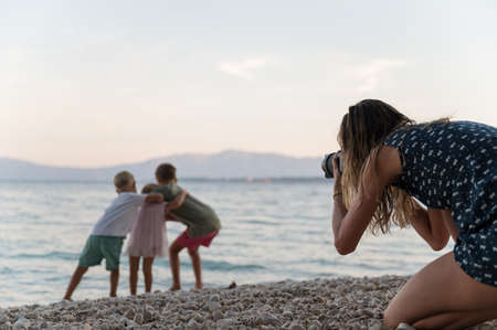 Mother taking photos of her three children hugging and playing on pebble beach at sunset.の写真素材