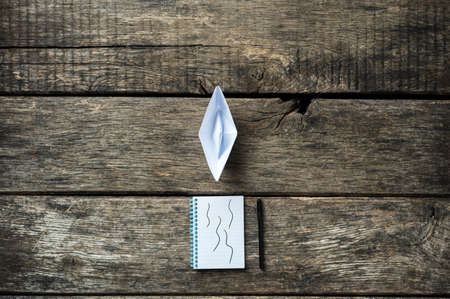 Notepad with a pen and paper made origami boat placed on wooden desk. Conceptual image of vision and...