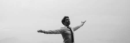 Greyscale wide view image of businessman standing under cloudy sky with his arms spread widely.の写真素材