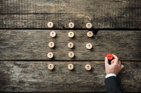 Hand of a businessman turning a red wooden cut circle with an arrow on it to point into a different direction than a group of others.の写真素材