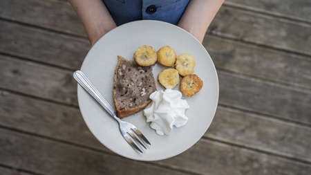 Top view of a child holding plate with tasty vegan dessert of home made banana bread, fried bananas and whipped cream.の写真素材