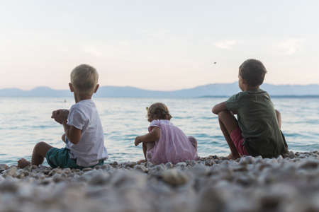 View from behind of three siblings, two boys and a girl, sitting on pebble beach by the sea in the evening playing and throwing stones.の写真素材