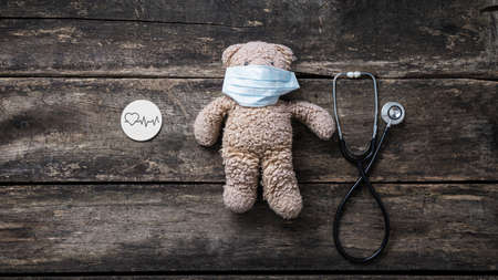 Top view of teddy bear wearing protective medical mask with stethoscope and  heartbeat tracing on wooden cut circle next to it. Over rustic wooden background.の写真素材