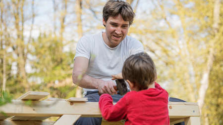 Young father building wooden playhouse with his toddler son outside in backyard.