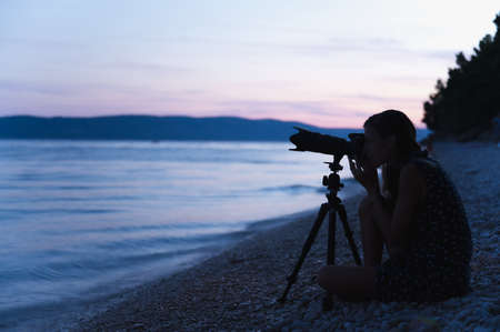 Young female photographer sitting on pebble beach in the evening with her camera set on tripod ready...