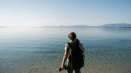 View from behind of a young male photographer holding his camera and tripod, standing by the beautiful calm sea water looking for a perfect spot to shoot.の写真素材