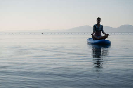 Peaceful young woman sitting in lotus position on sup board floating on calm sea water in early...