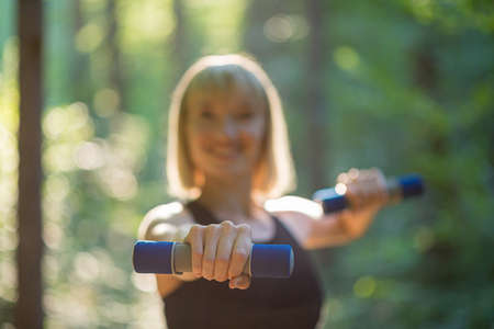 Young woman exercising with small hand weights outside in beautiful green forest. Focus to the hand.の写真素材
