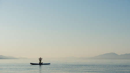 Young woman on floating sup board sitting in lotus position with her palm joined above her head.の写真素材