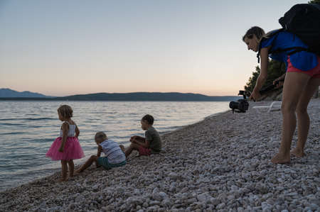 Young mother using a gimbal holder to video shoot her three kids playing at dusk on pebble beach by...