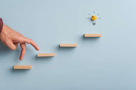 Male hand walking his fingers up the steps towards a glowing light bulb in a conceptual image of...