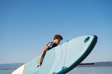 Young man carrying blue sup board out of the water after finishing his morning exercise.の写真素材