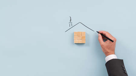 Male hand in a suit drawing a roof over a house build of wooden blocks in a conceptual image of real...