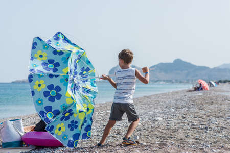 Toddler boy standing on beautiful pebble beach holding tight to sun umbrella as the wind in turning it over.の写真素材