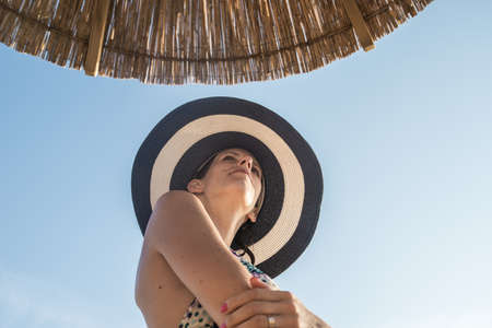 View from below of a young woman in bikini wearing sunhat standing under sun umbrella.