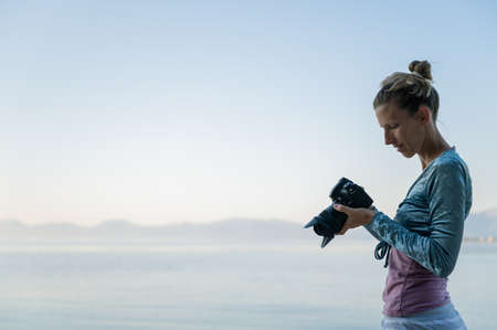 Young female photographer standing by the beautiful calm morning sea checking photos and setting on her professional dslr camera.の写真素材