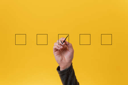 Male hand drawing a check mark in the middle box in a row of five. Over yellow background.の写真素材