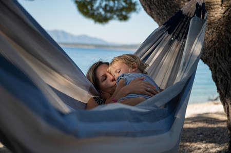 Young mother kissing her toddler daughter as they enjoy time together hanging in a hammock on the beach.の写真素材