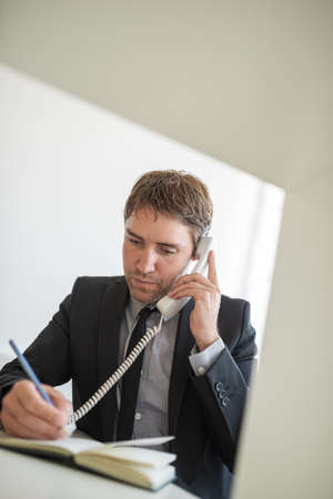 Handsome young businessman sitting at his office desk talking on white landline phone and making notes.の写真素材