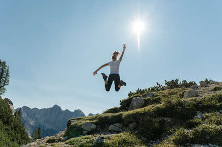View from behind of a young female hiker jumping up high enjoying life and the beautiful view of...