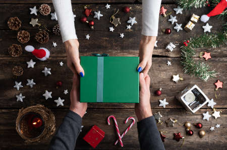 Top view of couples hands holding green holiday gift box over a rustic wooden desk full of christmas ornaments.の写真素材