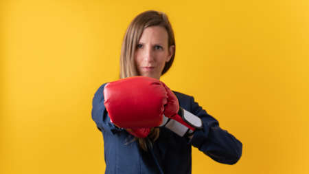 Confident young businesswoman wearing red boxing gloves punching towards the camera. Over yellow background.の写真素材