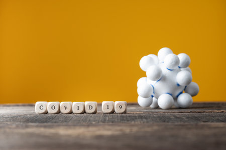 Covid 19 sign spelled on wooden dices next to a coronavirus molecule. Over yellow background.の写真素材