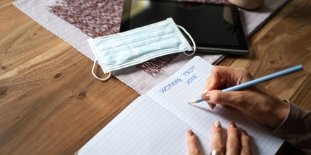 Working from home conceptual image - woman writing in notepad with digital tablet and medical protective mask on her home table.の写真素材