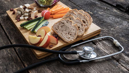 Healthy diet conceptual image - wooden serving plate full of vegetables, fruit sourdough bread and vegan protein next to a stethoscope.の写真素材