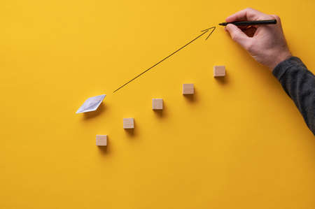 Male hand drawing an arrow pointing upwards in front of an origami made paper boat. Over yellow background.の写真素材