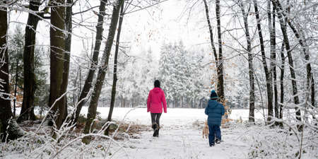 View from behind of a mother and her son walking in beautiful snowy forest with their cute small dog around them.の写真素材
