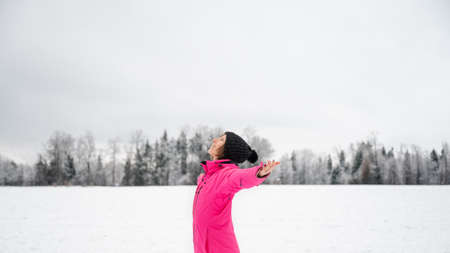Young woman in vibrant pink winter jacket standing in beautiful snowy nature with her arms spread widely meditating and enjoying peace.の写真素材