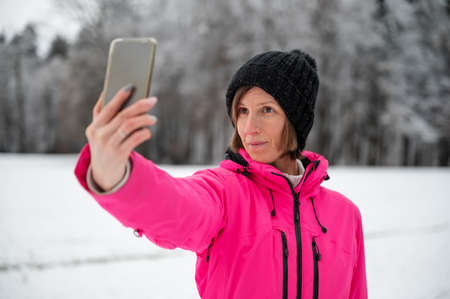 Young woman in pink winter jacket and black hat taking a selfie with her phone standing in beautiful snowy nature.の写真素材