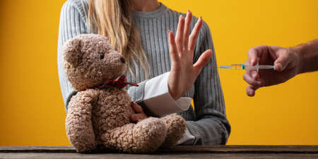 Woman making a stop gesture with her hand towards a vaccine while protecting her teddy bear in a conceptual image.の写真素材