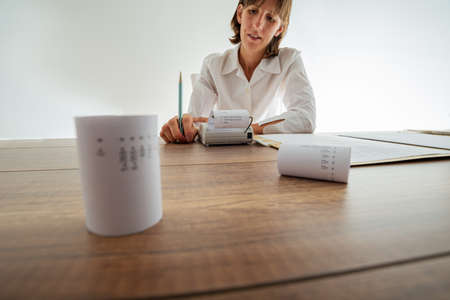 Young female accountant using adding machine working at her desk full of printout receipts and documents.の写真素材