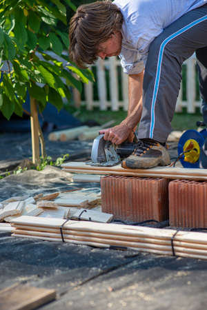 Young man working on a diy project outside in backyard sawing wooden planks for a fence.