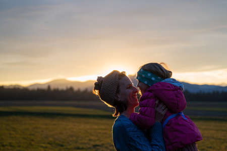 Cheerful smiling young mother lifting her toddler daughter while outside in a beautiful meadow at sunset.の写真素材