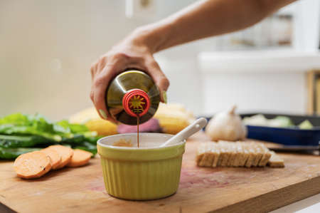 Closeup view of a woman pouring soya sauce into a cup to prepare a dip on wooden cutting board full of vegetables and vegan protein.の写真素材