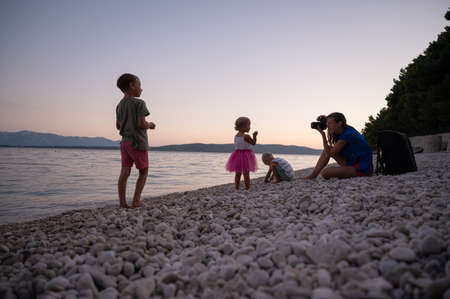 Young mother photographing her three children playing on pebble beach in a beautiful summer evening.の写真素材