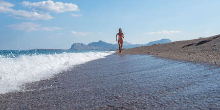 Young woman in pink bikini running on a beautiful pebble beach along the sea.の写真素材