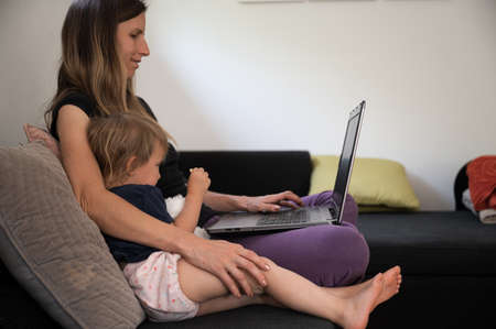 Young mother sitting on a sofa working on laptop computer with her toddler daughter cuddling next to...