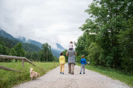 View from behind of a young father walking in green nature with his three kids, carrying one of them on his shoulders, with their dog alongside.の写真素材