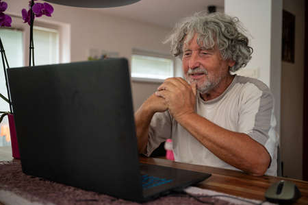 Senior man sitting at his dining table with a laptop computer in front of him talking to his family via video call.の写真素材
