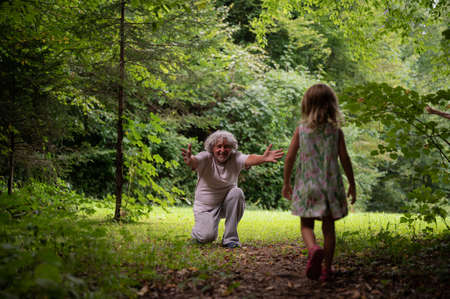 Grandpda kneeling down on the edge of a beautiful green meadow to greet his granddaughter with open arms.の写真素材