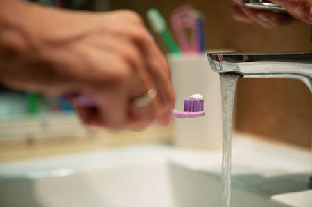 Closeup view of a hand holding toothbrush with toothpaste on it close to the tap water jet.の写真素材