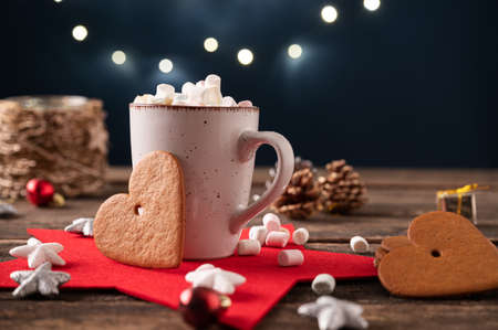 Hot cocoa topped with small marshmallows in a rustic cup placed on a wooden desk full of christmas holiday decorations and large heart shaped cookies.の写真素材